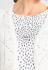 Cardigan bianco lavorato a maglia con motivo ad intaglio, sovrapposto a una maglietta bianca con design floreale a pois scuri.
