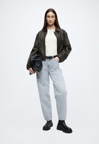 Blusão bomber de pele castanha, suéter creme, jeans largos azul claro e botas pretas até ao tornozelo. Modelo segura uma mala preta, exibindo um ajuste descontraído.