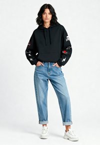 Felpa nera con cappuccio e disegni ricamati sulle maniche, abbinata a jeans azzurri arrotolati e sneakers bianche.