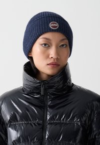 Caciula tricotată de culoare navy cu textură ribbed și un logo circular Colmar. Geacă puffer neagră cu luciu puternic, guler înalt și închidere cu fermoar.