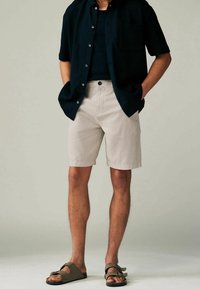 Homme portant un short beige arrivant aux genoux, une chemise noire à manches courtes avec des boutons, un t-shirt sombre en dessous, et des sandales vertes à bouts ouverts, debout avec les mains dans les poches.