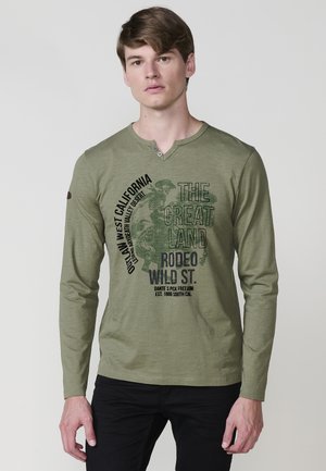Langärmliges olivgrünes T-Shirt mit V-Ausschnitt, das schwarze grafische Texte und Muster sowie Knöpfe am Ausschnitt aufweist. Schwarze Hose sichtbar.
