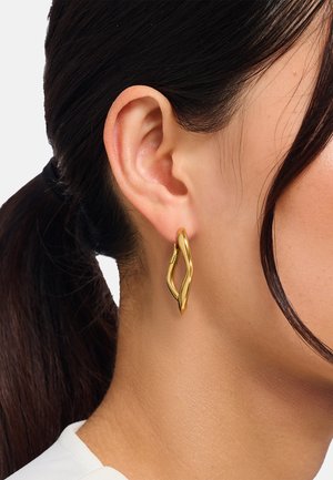 Goldene Creolen mit einem wellenförmigen, abstrakten Design. Glatte Oberfläche und glänzende Textur verstärken ihre optische Wirkung. Am Ohr befestigt.