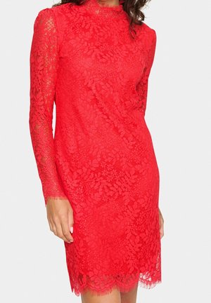 Femme portant une robe rouge en dentelle ajustée, à manches longues, avec des motifs floraux et un ourlet festonné, debout devant un fond uni.