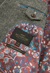 Veste en tissu pied-de-coq avec une poche doublée de bleu et une doublure intérieure à motifs floraux en bordeaux, comportant une étiquette pour la taille et l'ajustement.