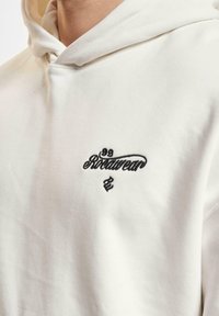 Hvid pullover hoodie lavet af blød bomuld. Har et sort broderet logo på brystet og en hætte med snøre. Glat tekstur.