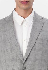 Blazer grigio a quadri con collo appuntito, indossato sopra una camicia bianca con bottoni. Il blazer presenta una chiusura a un bottone e un tessuto texturizzato.