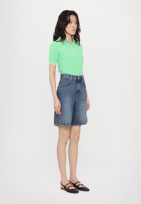Light green short-sleeve polo shirt with a button placket and white collar, paired with dark denim Bermuda shorts. Brown flat shoes.