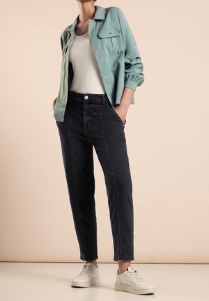 Personne portant une chemise verte claire boutonnée par-dessus un haut blanc, un jean noir taille haute et des baskets blanches, debout les mains dans les poches.