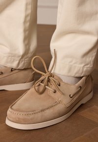 Beige Wildlederschuhe mit einer runden Zehenpartie, Gummisohle und Schnürverschluss. Getragen mit creamfarbener Hose, die eine sanfte Textur und Nähte zeigt.