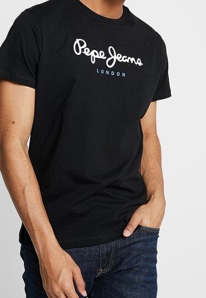 Mann trägt ein schwarzes Pepe Jeans London T-Shirt und dunkelblaue Jeans, mit einer Hand in der Tasche und der anderen locker an der Seite.