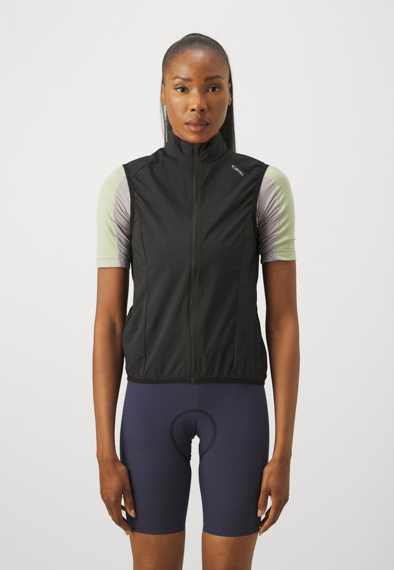Gilet de cyclisme sans manches noir avec un col haut, en tissu léger, et fermeture éclair à l'avant, superposé à un haut à manches courtes vert clair et un short bleu marine.