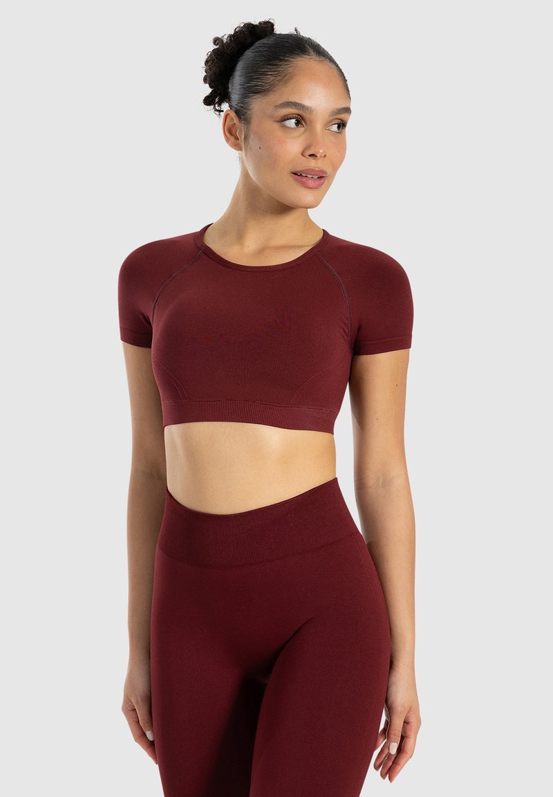 Mørkerød træningssæt bestående af en kortærmet crop top og højtaljede leggings, lavet af et glat, strækbart stof med dekorerende syninger.