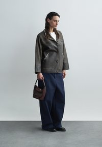 Bruine leren jas met zilveren studs, voorzakken, wijde blauwe broek en een kastanjebruine handtas. Het model staat tegen een neutrale achtergrond.