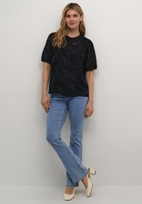 Blusa nera a maniche corte con design floreale texturizzato e chiusura a bottoni. Abbinata a jeans a campana di colore blu chiaro e scarpe con tacco di colore crema.