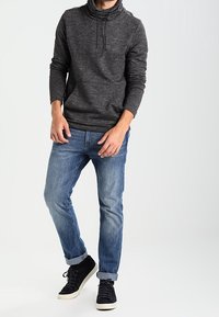 Man med en mörkgrå hoodie, uppkavlade blåjeans och svarta sneakers, som står mot en enkel ljus bakgrund.