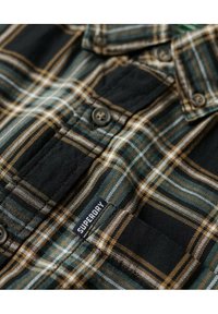 Camisa de cuadros verde y negra con rayas marrones y blancas, diseño de botones, textura de tela y una notable etiqueta de "Superdry".