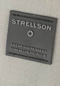 Etichetta in gomma nera con testo in rilievo "STRELLSON" e hashtag "#WEARINDEPENDENT." Coordinate stampate sotto su uno sfondo grigio testurizzato.