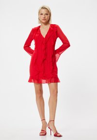 Rotes, halbtransparentes Kleid mit Rüschendetails an der Vorderseite und den Ärmeln, und einem mehrlagigen Rock. Kombiniert mit passenden Riemchenschuhen.