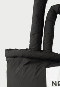 Sac tote isolé noir avec des poignées rembourrées, un fond plat, et une étiquette blanche avec du texte noir sur le côté. Texture lisse et mate.