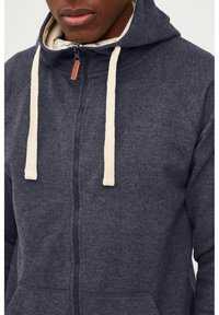 Sudadera con cremallera de color azul marino, hecha de tejido texturizado, con cordones de color crema y un bolsillo frontal. Cremallera con una lengüeta de cuero.