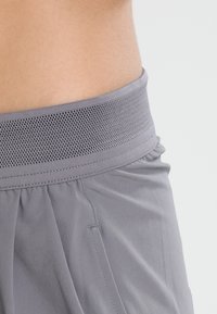 Gros plan sur la taille d’une personne portant un pantalon gris avec une large ceinture en mesh respirant sur un fond uni.