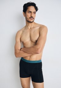Black boxer briefs with a teal waistband featuring "BJÖRN BORG" logo. Smooth texture, fitted design, and mid-thigh length.