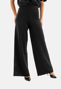 Pantaloni neri a gamba larga realizzati in materiale morbido, con un'alta vita e una texture liscia. Abbinati a tacchi a punta neri.