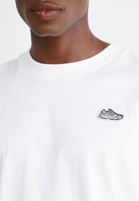 Man wearing a white shirt with a small embroidered sneaker patch on the left chest against a plain background.