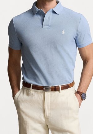 Mann in hellblauem Poloshirt mit Kragen, beigen Hosen, braunem Ledergürtel und braunem Armbanduhr mit blauen Zifferblatt, Hände in den Taschen.