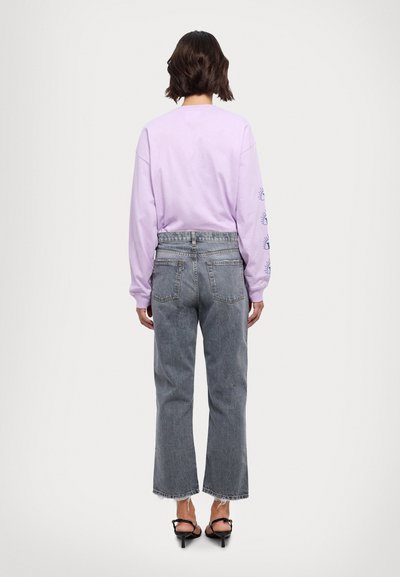 Camisa de manga larga lila con un gráfico azul en las mangas, combinada con unos jeans de mezclilla gris claro de cintura alta con dobladillos deshilachados. Sandalias negras de tacón.