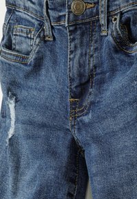 Los jeans de mezclilla azul presentan un aspecto desgastado con detalles de desgarros, costuras de color amarillo dorado y dos bolsillos delanteros con cierre de botón.