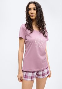 Moteris su ilgais garbanotais plaukais, vilkinti rožinę trumpomis rankovėmis marškinėlį su gėlių raštu ir užrašu „time to bloom“, derinant su rožiniais languotais šortais.