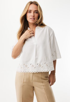 Blusa blanca de algodón con mangas cortas, con un dobladillo bordado en forma de ondas y delicados patrones florales. Combinada con pantalones beige.