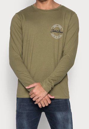 Man draagt een olijfgroen shirt met lange mouwen met het logo "Jack & Jones Originals" op de borst, gecombineerd met donkerblauwe jeans, armen gekruist.