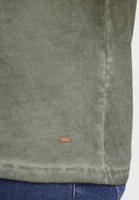 Olivengrøn bomulds T-shirt med en blød tekstur, der har et lille bronzefarvet mærke mærket "ISOLID" nær sømmen.