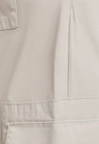 Světlý béžový materiál s hladkou texturou, se bočním švem a náprsní kapsou s viditelnými švy.