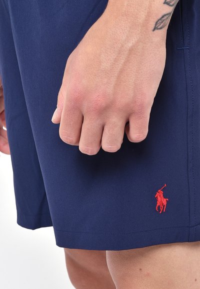 Pantaloni corti blu navy realizzati in tessuto liscio, con un logo ricamato rosso sull'orlo e una leggera lucentezza. Un primo piano di una mano che afferra il tessuto.