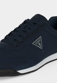 Baskets Guess bleu marine avec lacets noirs, semelle blanche et logo Guess triangulaire sur le côté.