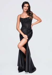 Femme en longue robe de soirée noire avec un corsage perlé et une fente haute sur la cuisse, posant avec une main sur la hanche et l'autre sur la jambe.