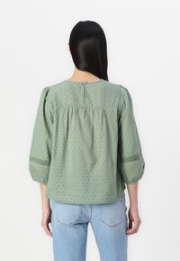 Blusa verde in cotone leggero con maniche a sbuffo e cuciture decorative. Collo rotondo con chiusura a bottone sul retro.