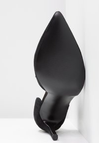Chaussure à talon haut noire à bout pointu avec semelle texturée, debout droite contre un coin blanc de mur et de sol.
