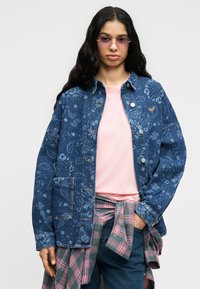 Sieviete ar gariem tumšiem matiem, valkājot rozā saulesbrilles, rozā krekliņu, zilu džinsa jaku ar rakstu un apakšā apjoztu svītrainu kreklu.