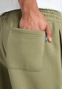 Olijfgroene sweaterbroek met een gestructureerde afwerking, voorzien van een grote zijzak en een elastische tailleband. De stof lijkt zacht en comfortabel te zijn.
