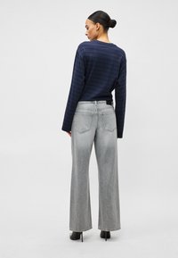 Moteris, turinti šukuoseną kuode, dėvi jūrinės mėlynos spalvos dryžuotą ilgomis rankovėmis palaidinę, šviesiai pilkus platėjančius džinsus ir juodus aukštakulnius batus, stovėdama nugara.