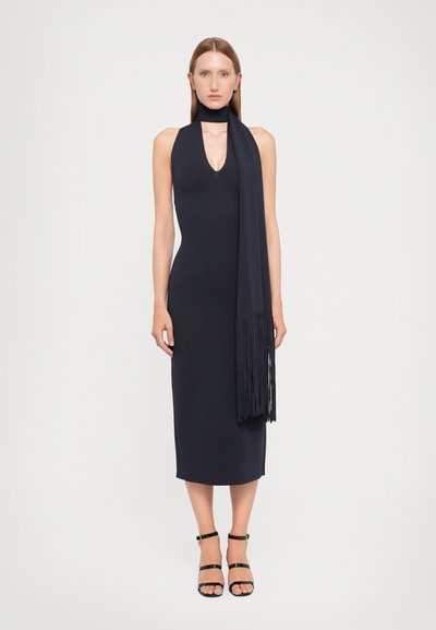 Vestito senza maniche di colore blu navy con scollatura a V profonda e una lunga sciarpa con frange. Silhouette aderente e design fino al ginocchio, abbinato a sandali neri.