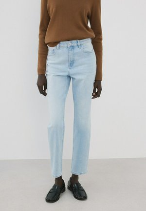 Persona con vaqueros de pierna recta azul claro, camiseta de manga larga marrón y mocasines negros, de pie contra un fondo liso y claro.