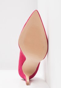 Chaussure à talon haut en daim rose à bout pointu, vue de la semelle avec une semelle extérieure beige striée et un talon fin, sur fond blanc.