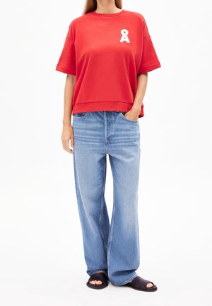 Vrouw draagt een rood T-shirt met korte mouwen en wit logo, losse lichtblauwe spijkerbroek en zwarte slides, staand met een hand in de zak.