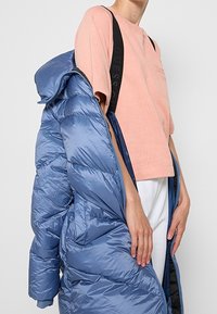 Une personne portant une chemise rose à manches courtes et un pantalon blanc tient un long manteau en duvet bleu sur une épaule.